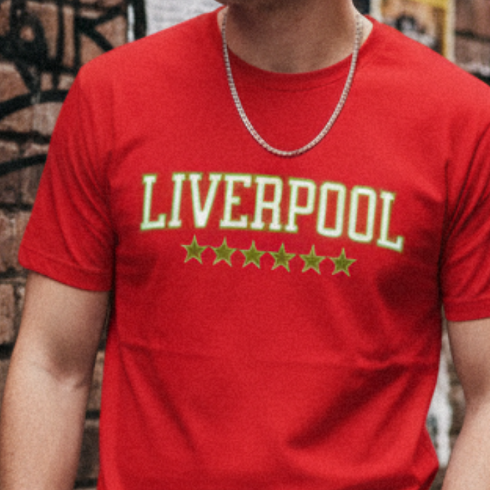 Person wearing a red 'Liverpool' t-shirt with five stars against a brick wall.