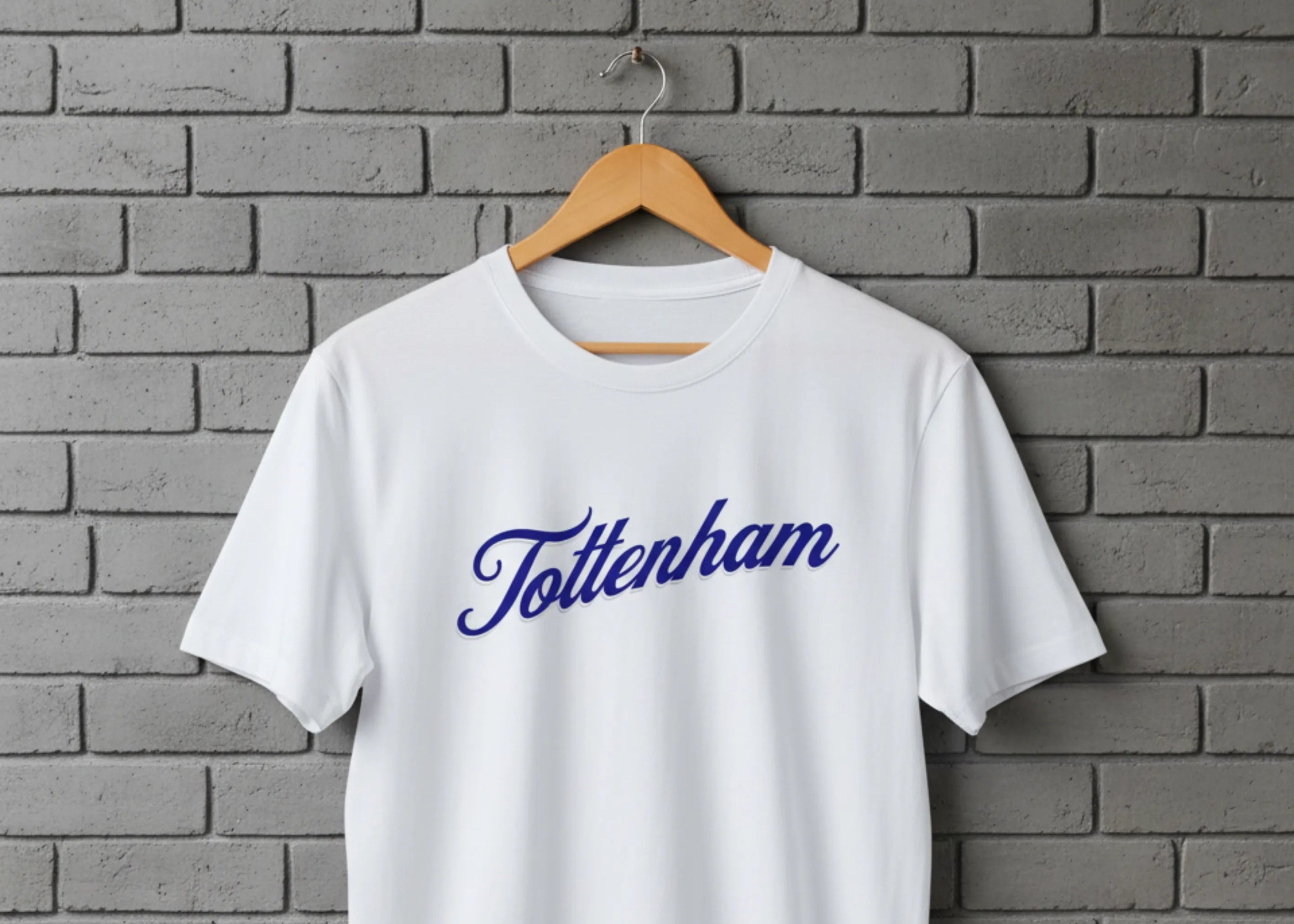 White t-shirt with 'Tottenham' printed on it, hanging against a gray brick wall.