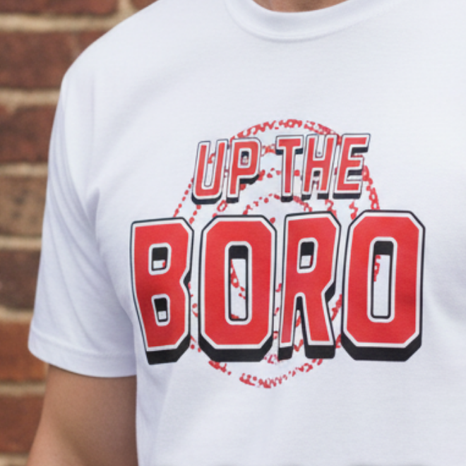 White t-shirt with 'Up the Boro' design, displayed with other colored shirts against a brick wall.