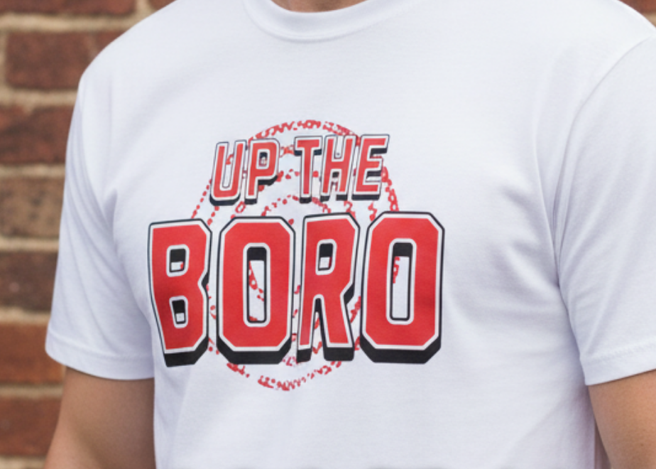 White t-shirt with 'Up the Boro' design, displayed with other colored shirts against a brick wall.